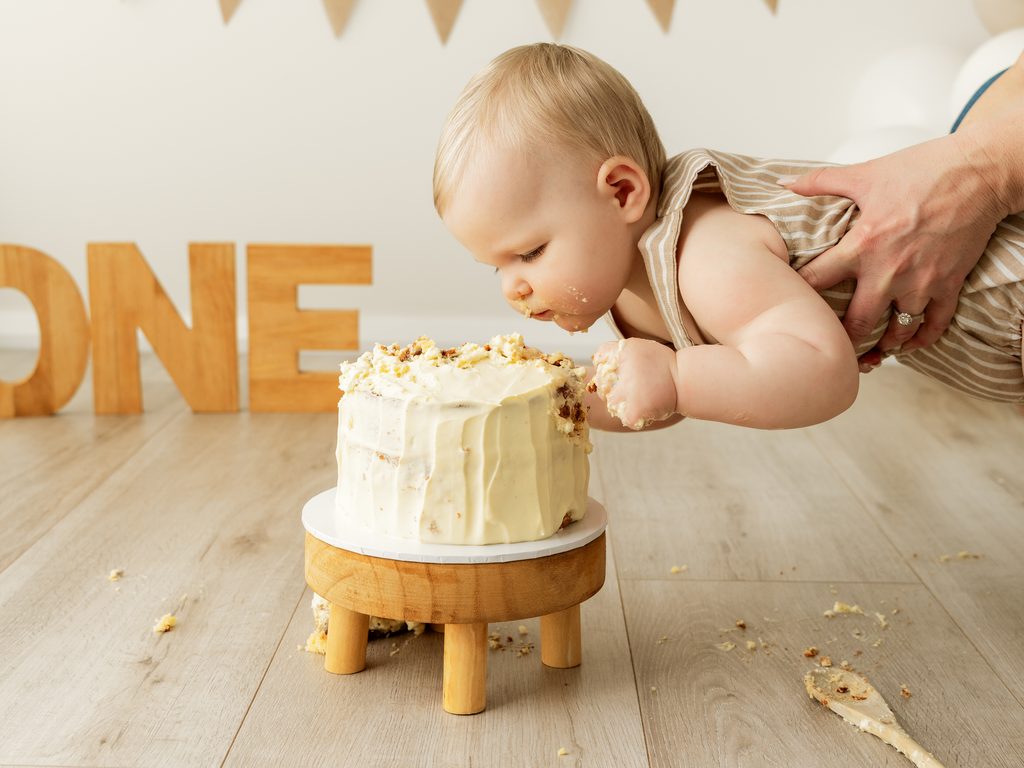 Baby leaning forward to explore a white birthday cake during a first birthday cake smash session.