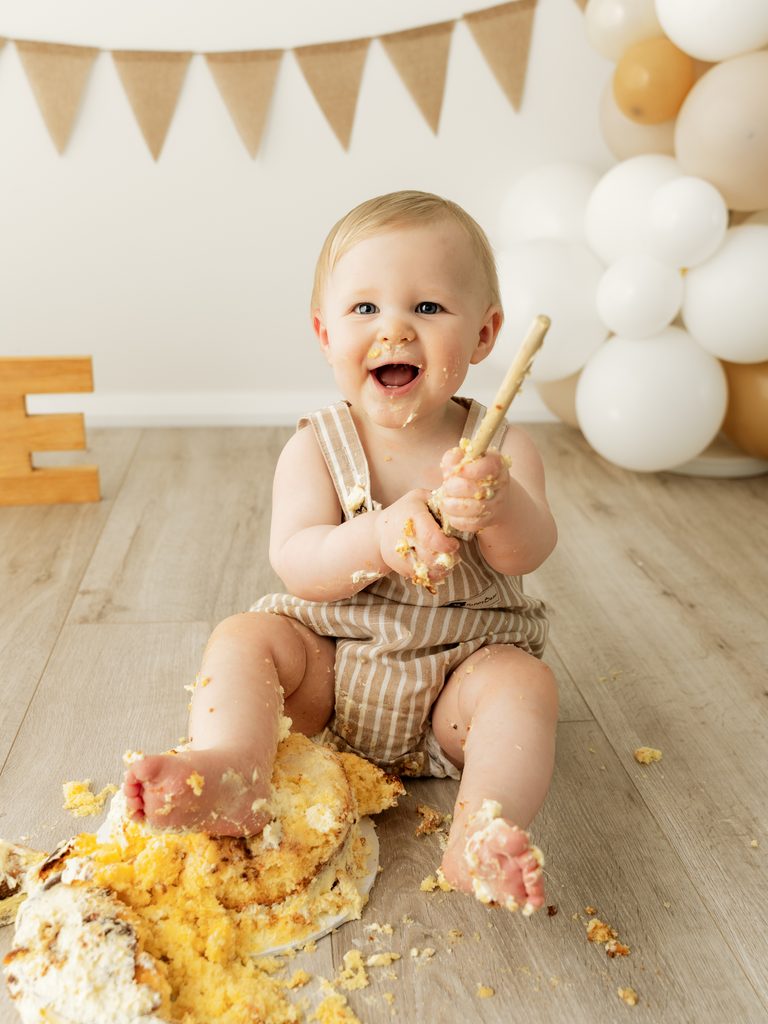 Happy one-year-old baby sitting among pieces of smashed birthday cake with white balloons behind.