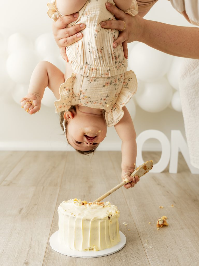 Playful upside-down portrait of a one-year-old baby holding a wooden spoon above their birthday cake.