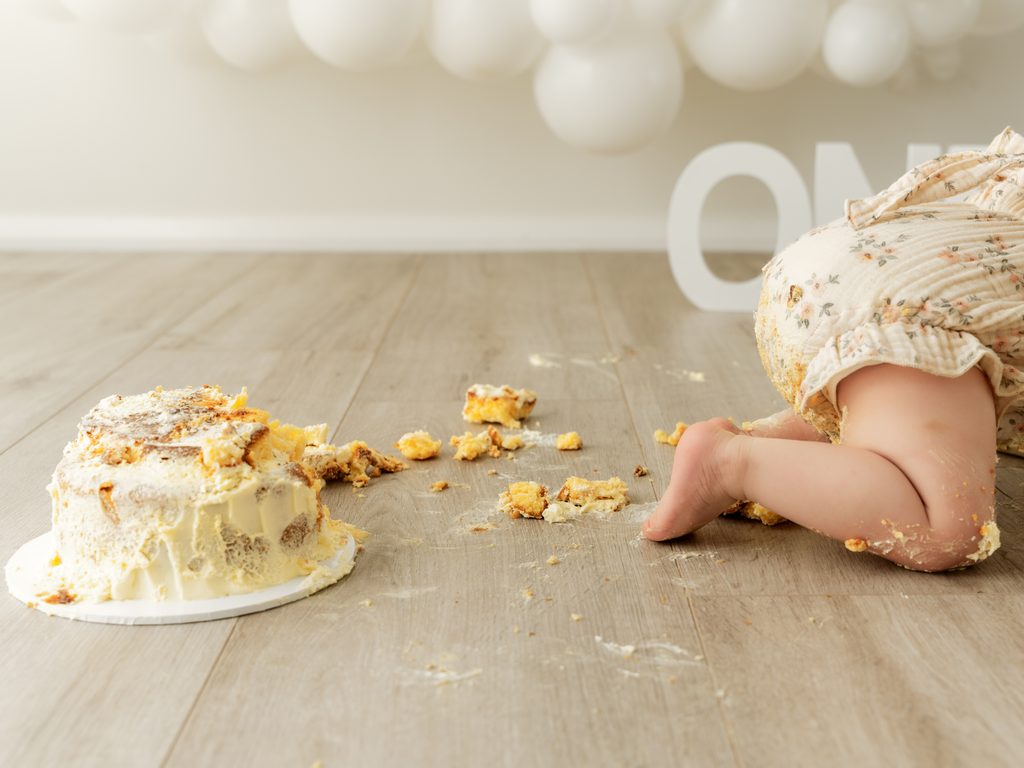 Smashed birthday cake scattered across the studio floor beside a baby's little feet.