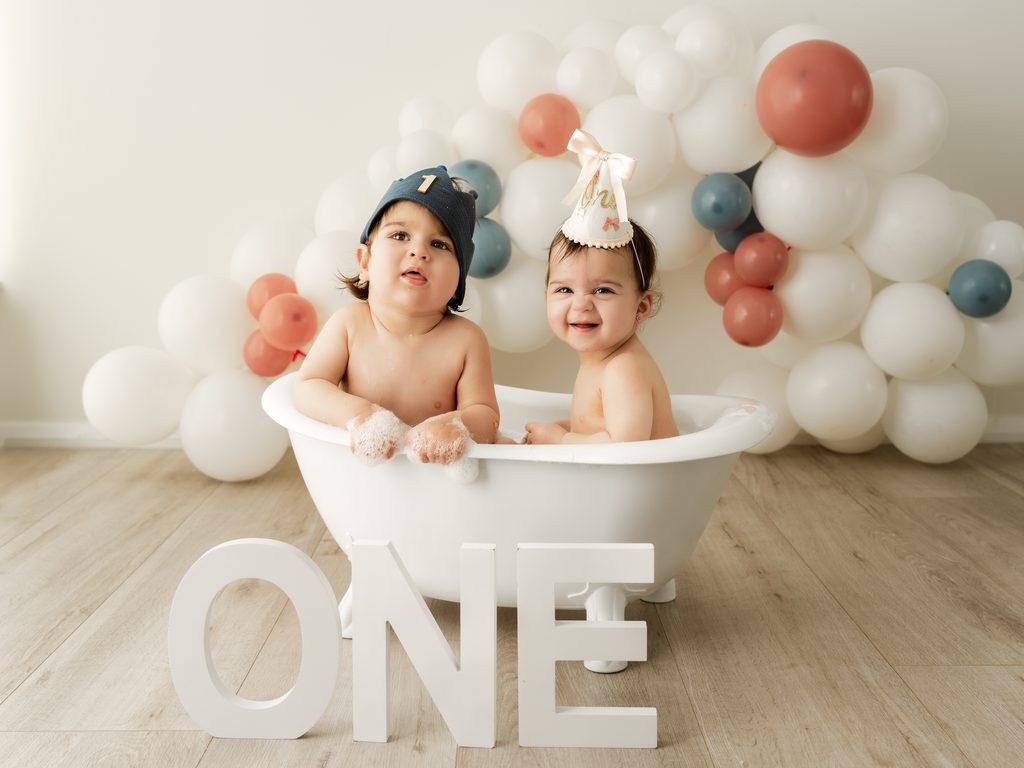 Baby splashing in a small white bathtub during a first birthday photography session.
